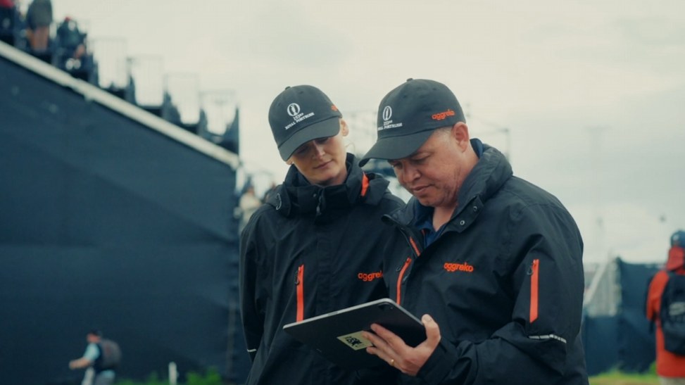 Frame from the Aggreko end of year wrap: two Aggreko crew in branded jackets checking a tablet on site at a golf championship.